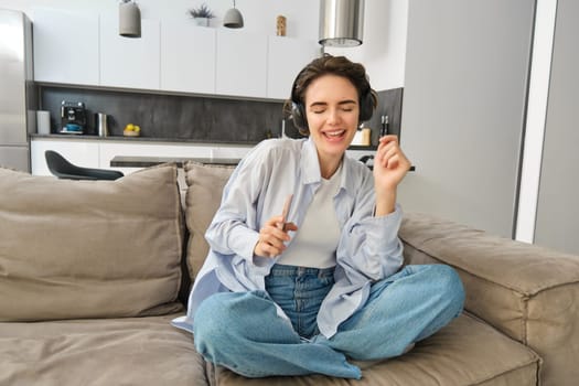 Portrait of happy girl in headphones, dancing on sofa and listening music, having fun at her house.