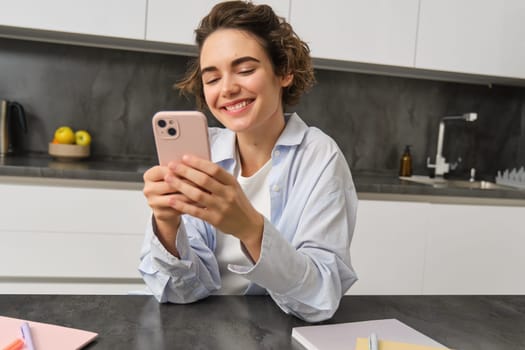 Technology and lifestyle. Young woman sits at home, uses smartphone in her kitchen and smiles.