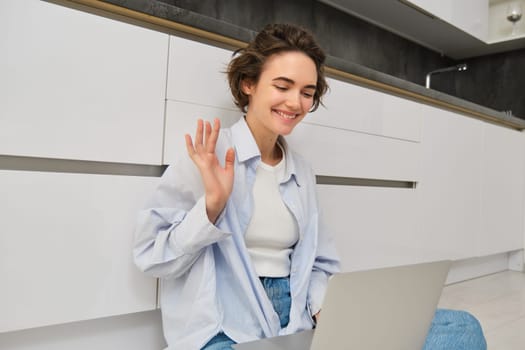 Friendly girl talks on laptop, connects to work online meeting via computer, sits on floor and waves hand at camera.