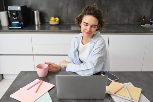 Beautiful brunette girl, student studying online course from home, tutor working on remote, looking at laptop, sitting in kitchen with cup of coffee.