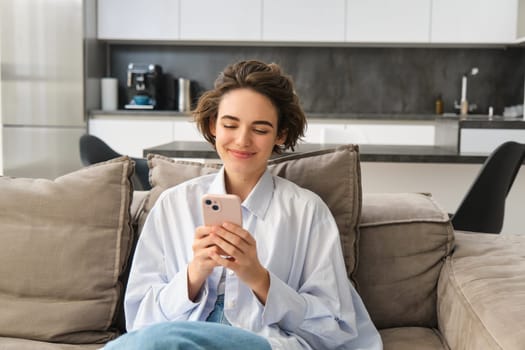 Image of happy woman sits on couch with smartphone, looks at mobile phone screen with pleased smile, chats in app, watches videos online.