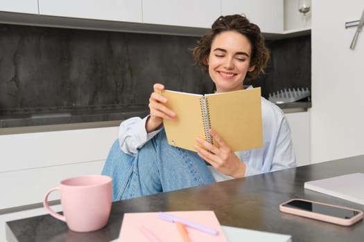 Smiling beautiful woman, sitting with notebook in kitchen, reading notes, studying, doing homework.