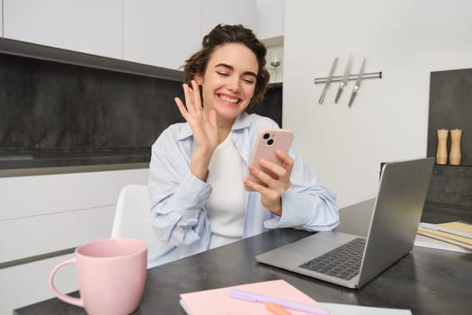 Happy young woman says hello, waves hand at smartphone and smiles, video chats with someone on mobile phone app.