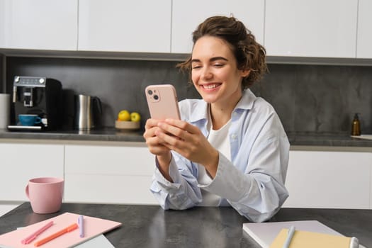 Technology and lifestyle. Young woman sits at home, uses smartphone in her kitchen and smiles.