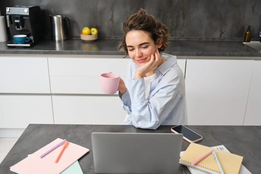 Beautiful brunette girl, student studying online course from home, tutor working on remote, looking at laptop, sitting in kitchen with cup of coffee.