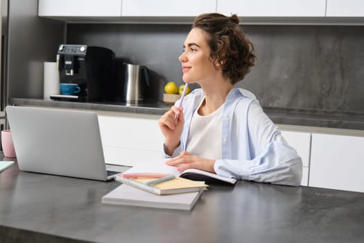 Portrait of young woman thinking, working on laptop at home, writing down, makes notes while does her homework, studies online course, listens to webinar.