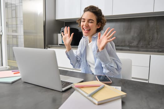 Beautiful girl waves hands at laptop camera, says hello to coworkers, connects to work meeting online, studies on remote from her kitchen.