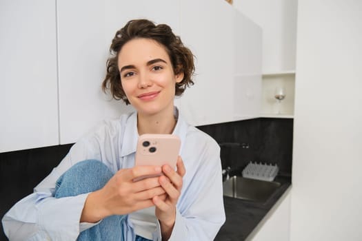 Portrait of smiling beautiful woman, sitting in kitchen with smartphone, looking happy at camera, spending time at home, using mobile phone app.