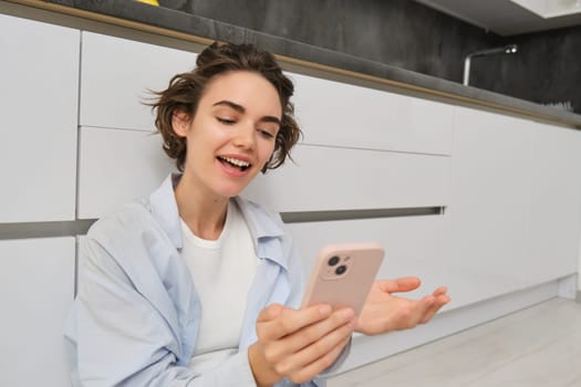 Portrait of happy young woman, shows her brand new kitchen, sits on floor, points at furniture and smiles, video chats on smartphone. Technology and lifestyle concept