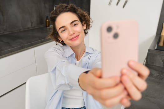 Beautiful girl takes selfie on smartphone in her kitchen, makes photos at home on mobile phone.