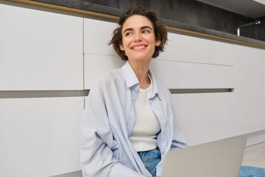 Freelance and lifestyle concept. Young woman sits on floor with laptop, works from home, sets up her remote workspace in kitchen, does homework.