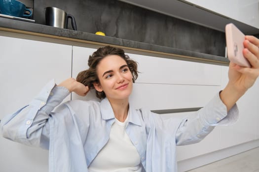 Beautiful young woman takes selfie on mobile phone, poses for photo on smartphone, smiles, sits on kitchen floor, creates content for her social media profile.