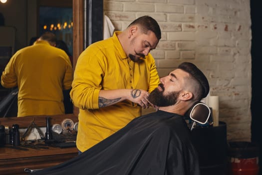 Client with luxurious black beard sit on chair, and professional barber make beard haircut in barber shop with trimmer