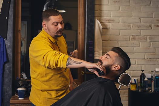 Client with luxurious black beard sit on chair, and professional barber make beard haircut in barber shop with trimmer