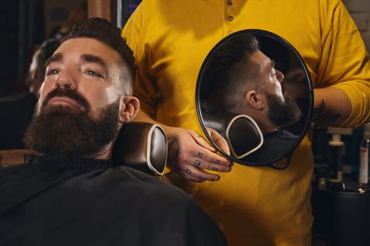 Client with luxurious black beard sit on chair, and professional barber make beard haircut in barber shop with trimmer