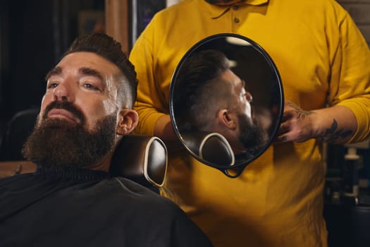 Client with luxurious black beard sit on chair, and professional barber make beard haircut in barber shop with trimmer