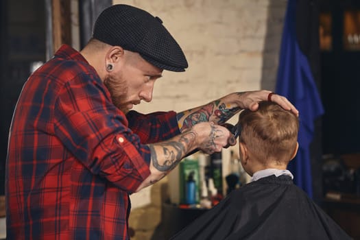 A pretty blonde boy happy to be on the haircut with a professional hairdresser. Blond little boy having a haircut at hair salon. Hairdresser's hands making hairstyle to child at barbershop