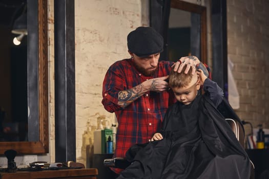 A pretty blonde boy happy to be on the haircut with a professional hairdresser. Blond little boy having a haircut at hair salon. Hairdresser's hands making hairstyle to child at barbershop