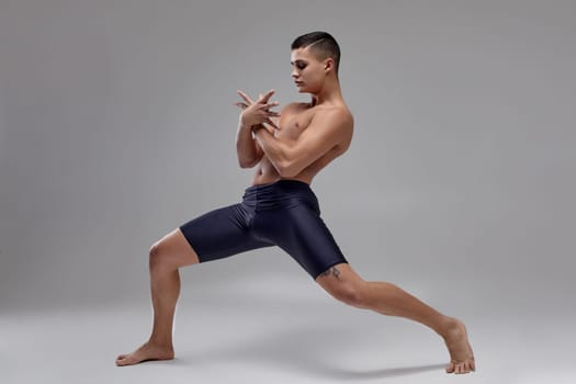 Full length portrait of an attractive muscular male ballet dancer, dressed in a black shorts. He is making a dance element with crossed hands, against a gray background in studio. Bare legs and torso. Ballet and contemporary choreography concept. Art photo.