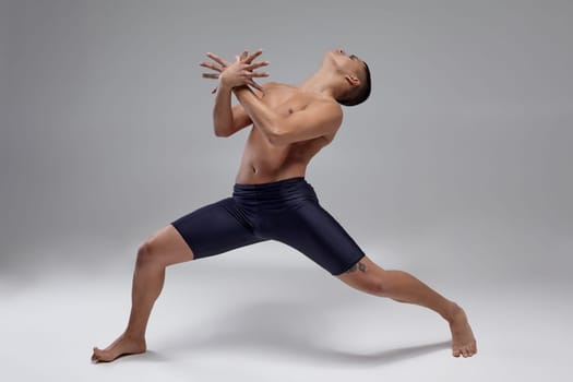 Full length portrait of a good-looking muscular male ballet dancer, dressed in a black shorts. He is making a dance element with crossed hands and looking up, against a gray background in studio. Bare legs and torso. Ballet and contemporary choreography concept. Art photo.