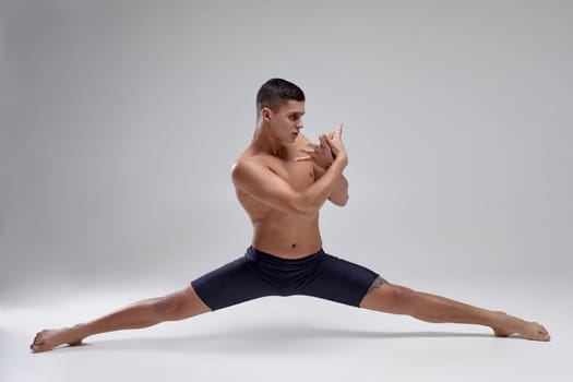 Full length photo of a good-looking muscular man ballet dancer, dressed in a black shorts. He is doing a twine against a gray background in studio. Bare legs and torso. Ballet and contemporary choreography concept. Art photo.