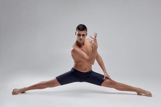 Full length portrait of a handsome muscular man ballet dancer, dressed in a black shorts. He is doing a twine against a gray background in studio. Bare legs and torso. Ballet and contemporary choreography concept. Art photo.