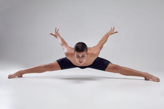 Portrait of a handsome man ballet dancer, dressed in a black shorts. He is doing a twine on a gray background in studio. Bare legs and torso. Ballet and contemporary choreography concept. Art photo.