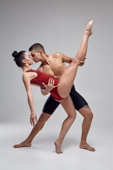 Couple of a young graceful ballet dancers are posing over a gray studio background. Man in black shorts and woman in a red swimwear are dancing together. Ballet and contemporary choreography concept. Art photo.