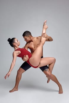 Two talanted athletic ballet dancers are posing over a gray studio background. Good-looking man in black shorts and charming woman in a red swimwear are dancing together. Ballet and contemporary choreography concept. Art photo.