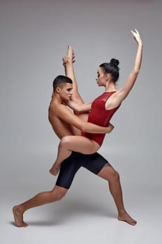 The pair of a graceful young ballet dancers are posing over a gray studio background. Strong man in black shorts and tiny woman in a red swimwear are dancing together. Ballet and contemporary choreography concept. Art photo.