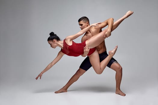 Pair of a graceful athletic ballet dancers are posing over a gray studio background. Handsome male in black shorts and charming female in a red swimwear are dancing together. Ballet and contemporary choreography concept. Art photo.