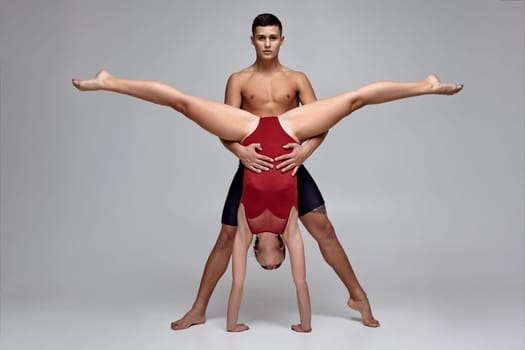 The pair of a graceful athletic ballet dancers are posing against a gray studio background. Good-looking man in black shorts and lovely woman in a red swimwear are dancing together. Ballet and contemporary choreography concept. Art photo.