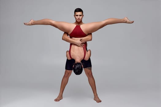 Pair of a modern ballet dancers are posing over a gray studio background. Handsome man in black shorts and beautiful girl in a red swimwear are dancing together. Ballet and contemporary choreography concept. Art photo.