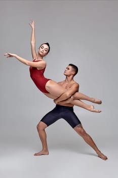 The couple of a young beautiful ballet dancers are posing over a gray studio background. Male in black shorts and female in a red swimwear are dancing together. Ballet and contemporary choreography concept. Art photo.