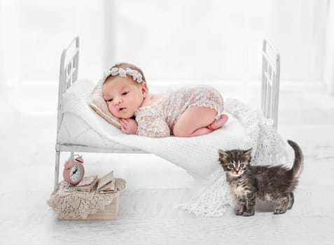 Beautiful newborn baby girl and kitty lying on her tummy on stylized white bed during studio photoshoot. Cute portrait of infant child in white room