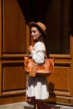 brunette with long hair wearing white dress and beige hat posing with luxury leather bag. wooden background. Vacation, tourism and travel