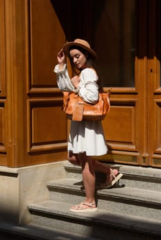 brunette with long hair wearing white dress and beige hat posing with luxury leather bag. wooden background. Vacation, tourism and travel