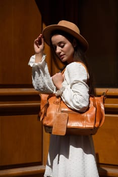 brunette with long hair wearing white dress and beige hat posing with luxury leather bag. wooden background. Vacation, tourism and travel