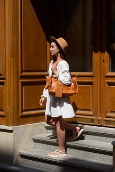 brunette with long hair wearing white dress and beige hat posing with luxury leather bag. wooden background. Vacation, tourism and travel