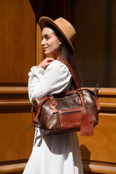 brunette with long hair wearing white dress and beige hat posing with luxury leather bag. wooden background. Vacation, tourism and travel