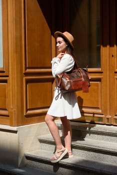 brunette with long hair wearing white dress and beige hat posing with luxury leather bag. wooden background. Vacation, tourism and travel