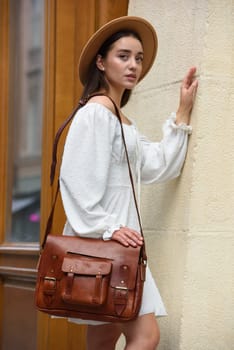 brunette with long hair wearing white dress and beige hat posing with luxury leather bag. wooden background. Vacation, tourism and travel