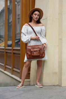 brunette with long hair wearing white dress and beige hat posing with luxury leather bag. wooden background. Vacation, tourism and travel