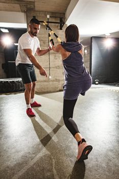 Rear view of young handsome woman having workout with a suspension straps at the gym supported of her personal male trainer. 