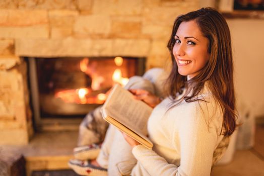 Beautiful young smiling woman reading book and enjoying by the fireplace.