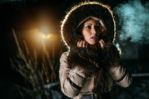 Beautiful young worried woman dressed in winter jacket with fur hood is standing in winter night and looking away waiting for somebody.
