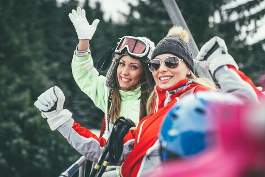 Beautiful young woman friends enjoying in winter vacations. They driving on ski lift and looking at camera with smile.