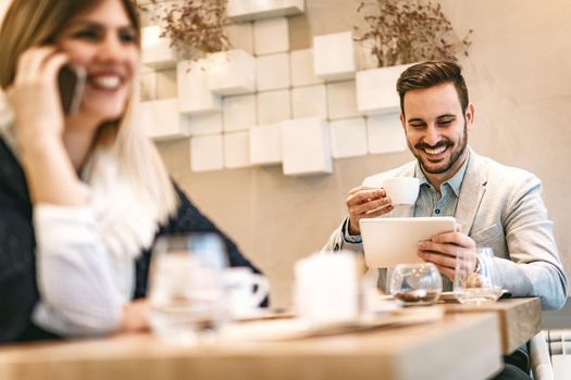 Young smiling businessman on a break in a cafe. He is checking social media at digital tablet and drinking coffee. 