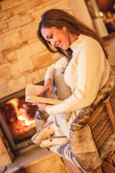 Beautiful young smiling woman reading book and enjoying by the fireplace.
