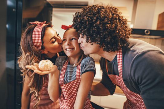 Happy parents and their daughter enjoy and having fun in making and having healthy meal together at their home kitchen. Parents are kissing a little girl while she is holding a mandarin ready to eat.